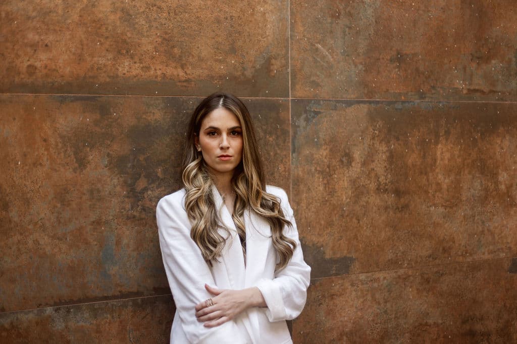 Lais Nohemann, fundadora da NÔH | BI, de blazer branco e braços cruzados, diante de uma parede terracota